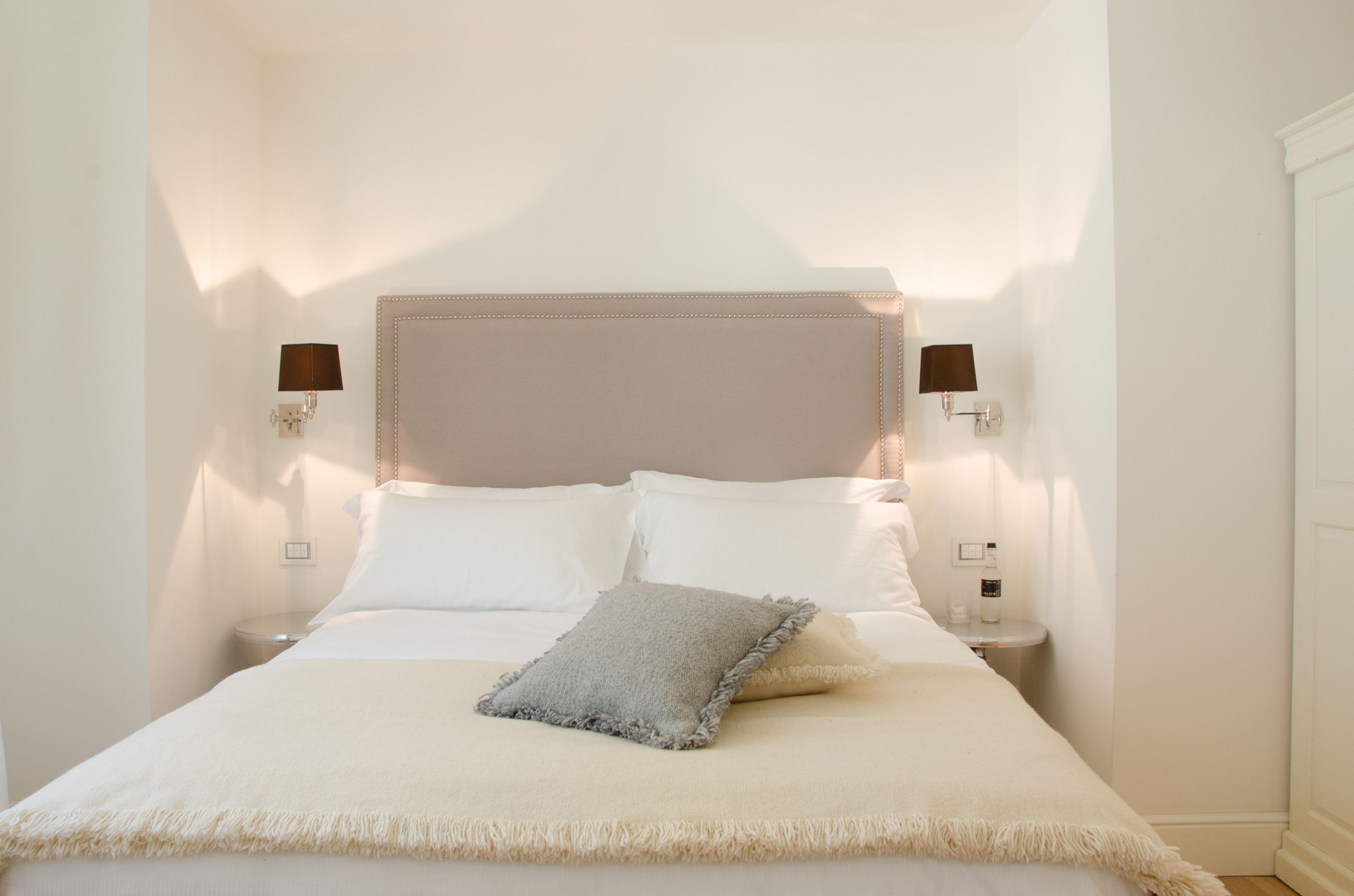 Un lit bien fait, avec une tête de lit beige, des oreillers blancs et un coussin décoratif gris. Deux lampes murales sont fixées de chaque côté du lit.