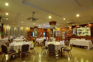 Uma sala de banquetes com mesas redondas dispostas para refeições, painéis de madeira e um espaço de bar.
