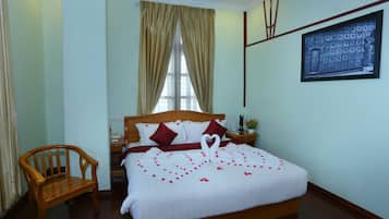 A neatly made bed with a heart-shaped decoration, a wooden chair, a nightstand with a lamp, and a framed picture on the wall.