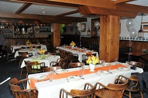 Un restaurant avec plusieurs tables dressées pour le service, des chaises en bois et un bar situé au fond de la salle.