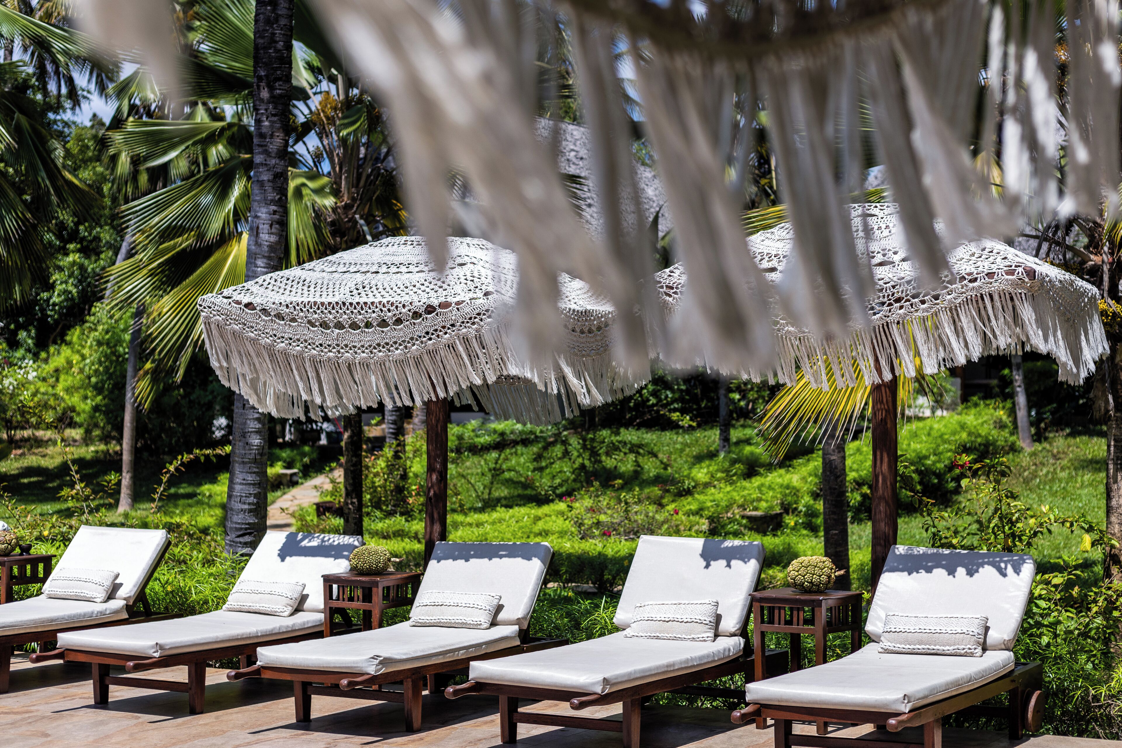 A row of wooden lounge chairs with white cushions and decorative pillows, set against a lush green garden with palm trees and a thatched umbrella.