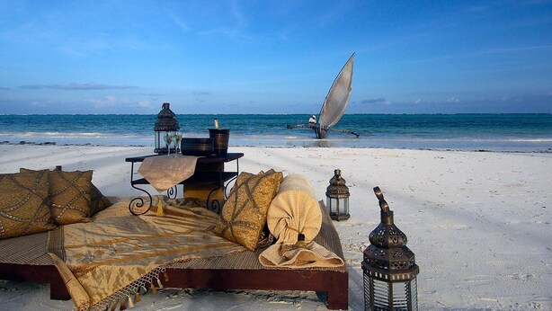 A beach setting with a bed, lanterns, and a sailboat in the background. - The Palms Zanzibar - All Inclusive (Dongwe)