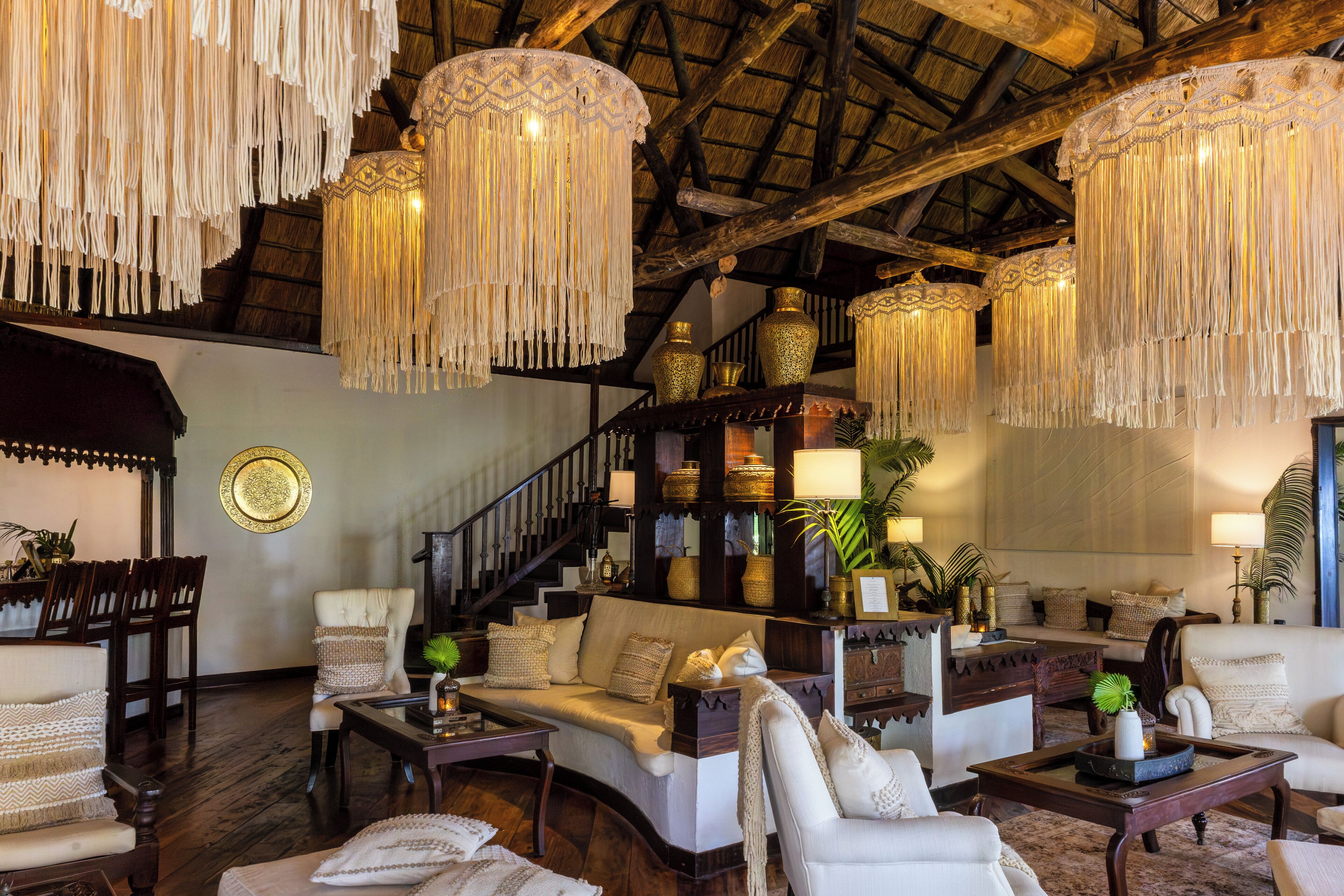 A spacious living area with a high ceiling, wooden beams, and a staircase. There are multiple seating areas with sofas and armchairs, and a variety of decorative items including vases and plants.