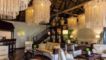A spacious living area with a high ceiling, wooden beams, and a staircase. There are multiple seating areas with sofas and armchairs, and a variety of decorative items including vases and plants.