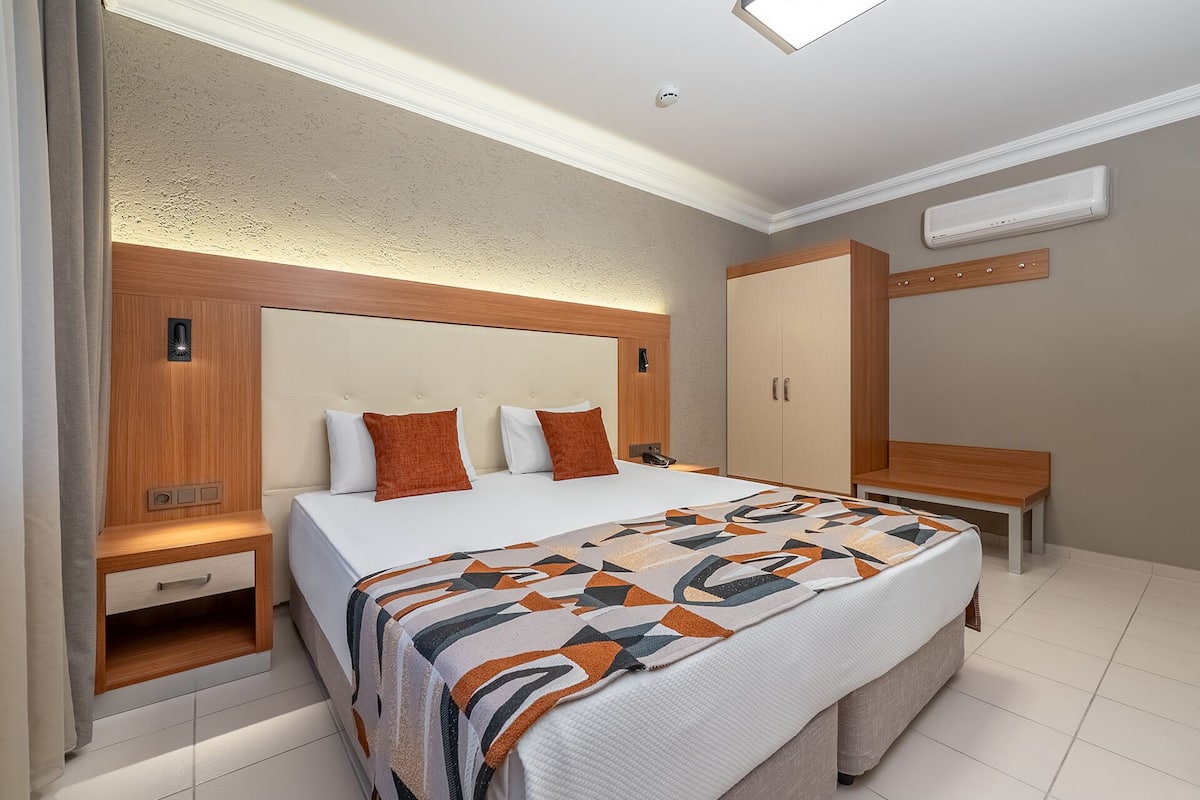 A modern hotel room with a large bed, a wooden headboard, a wardrobe, and a bedside table with a lamp.