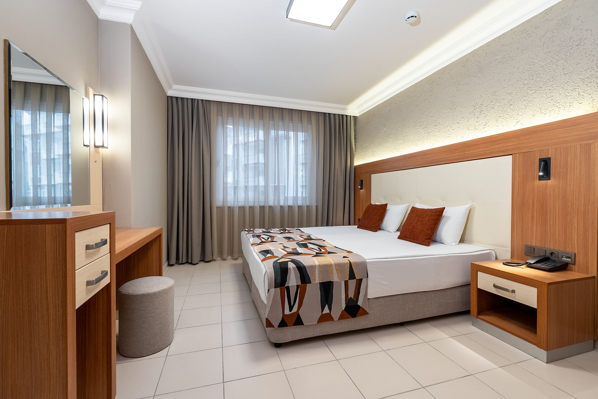 A modern hotel room with a large bed, a wooden nightstand, a mirror, and a window with curtains.