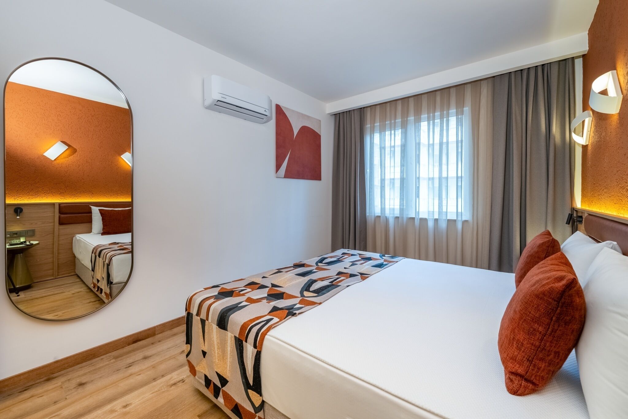 A hotel room with a bed, a large mirror, an air conditioning unit, a window with curtains, and a wall-mounted light fixture.