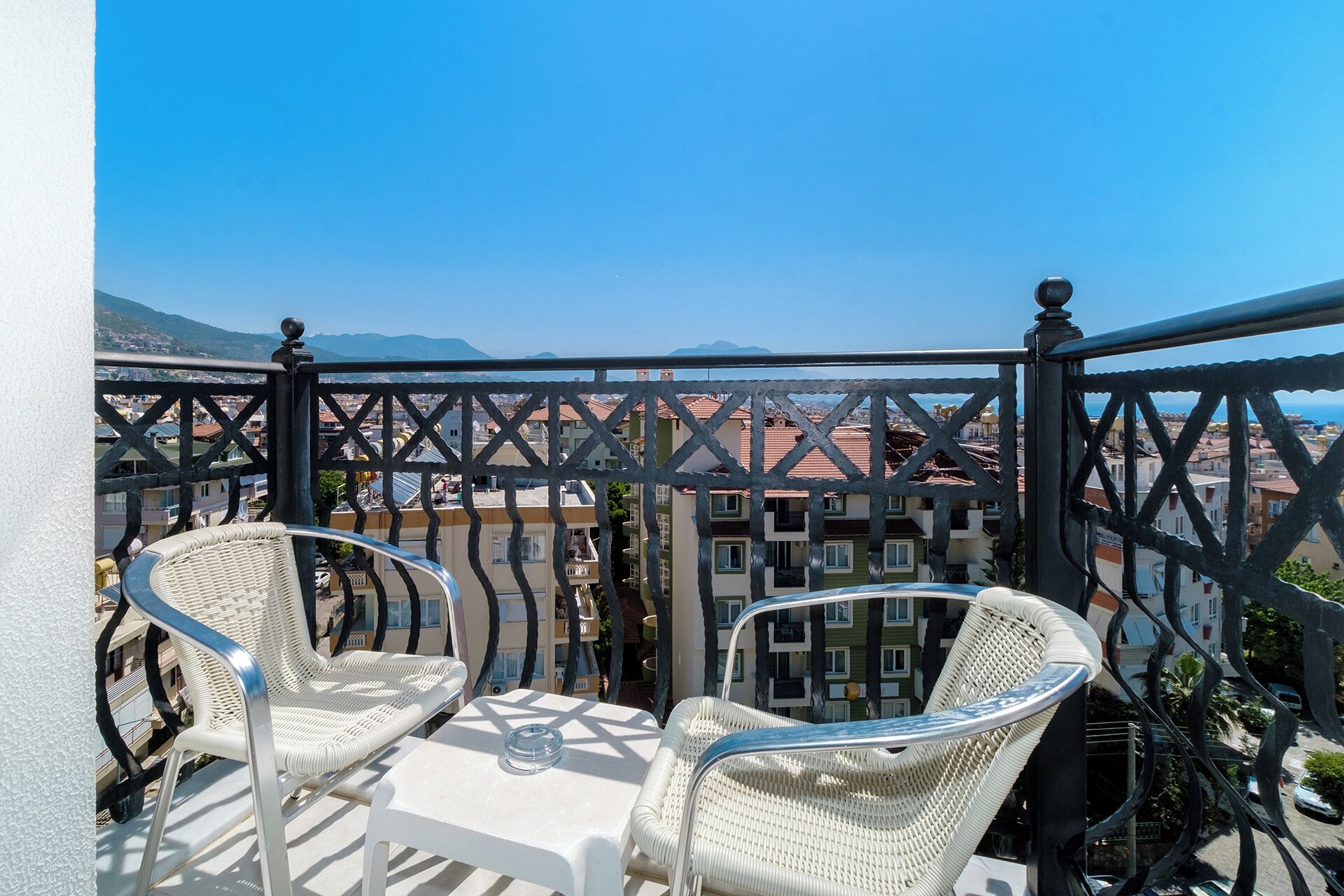 Un balcon avec deux fauteuils en osier et une table, d’où l’on a vue sur une ville côtière, avec des bâtiments et des montagnes en arrière-plan.