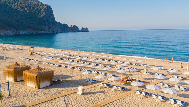 A beach with rows of sun loungers and umbrellas, a lifeguard tower, and a mountain in the background. - Villa Sunflower Hotel (Alanya)