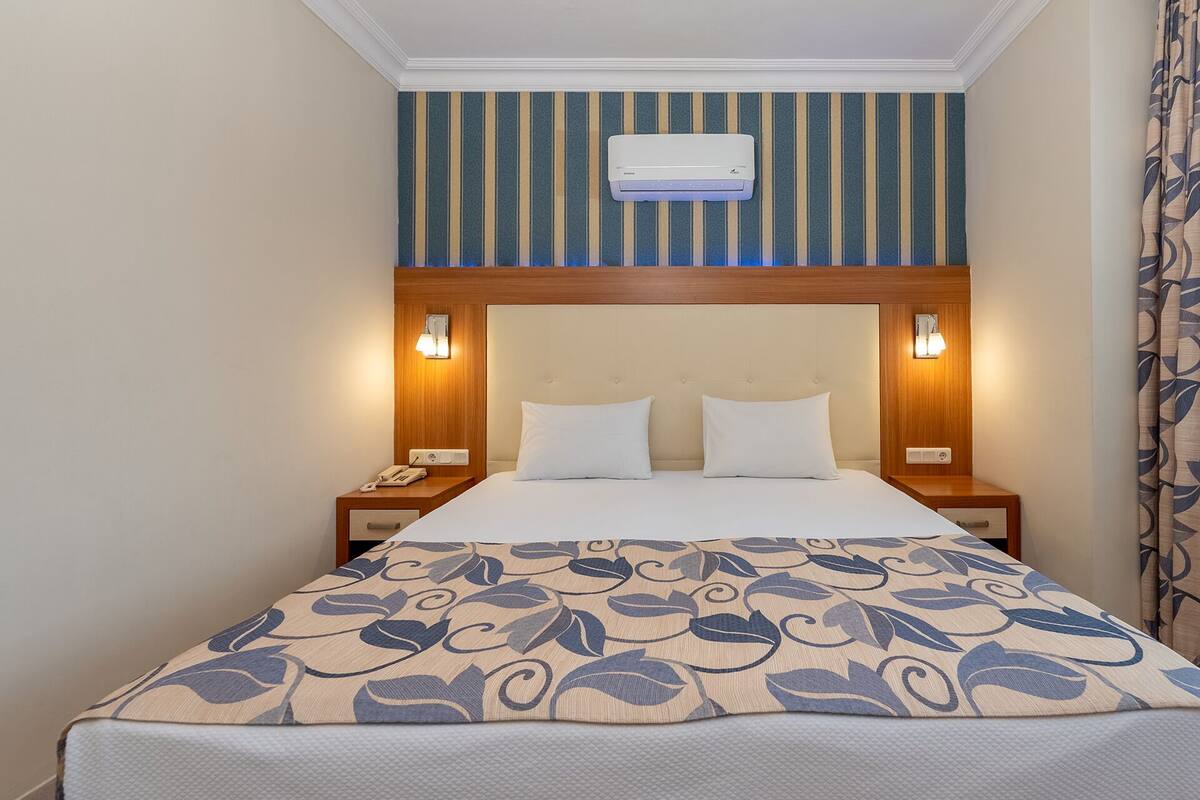 A hotel room with a bed, two bedside tables, a wall-mounted air conditioner, and a striped wall.