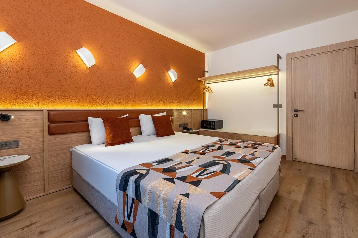 A modern hotel room with a large bed, a bedside table, a nightstand, a wall-mounted lamp, and a door.
