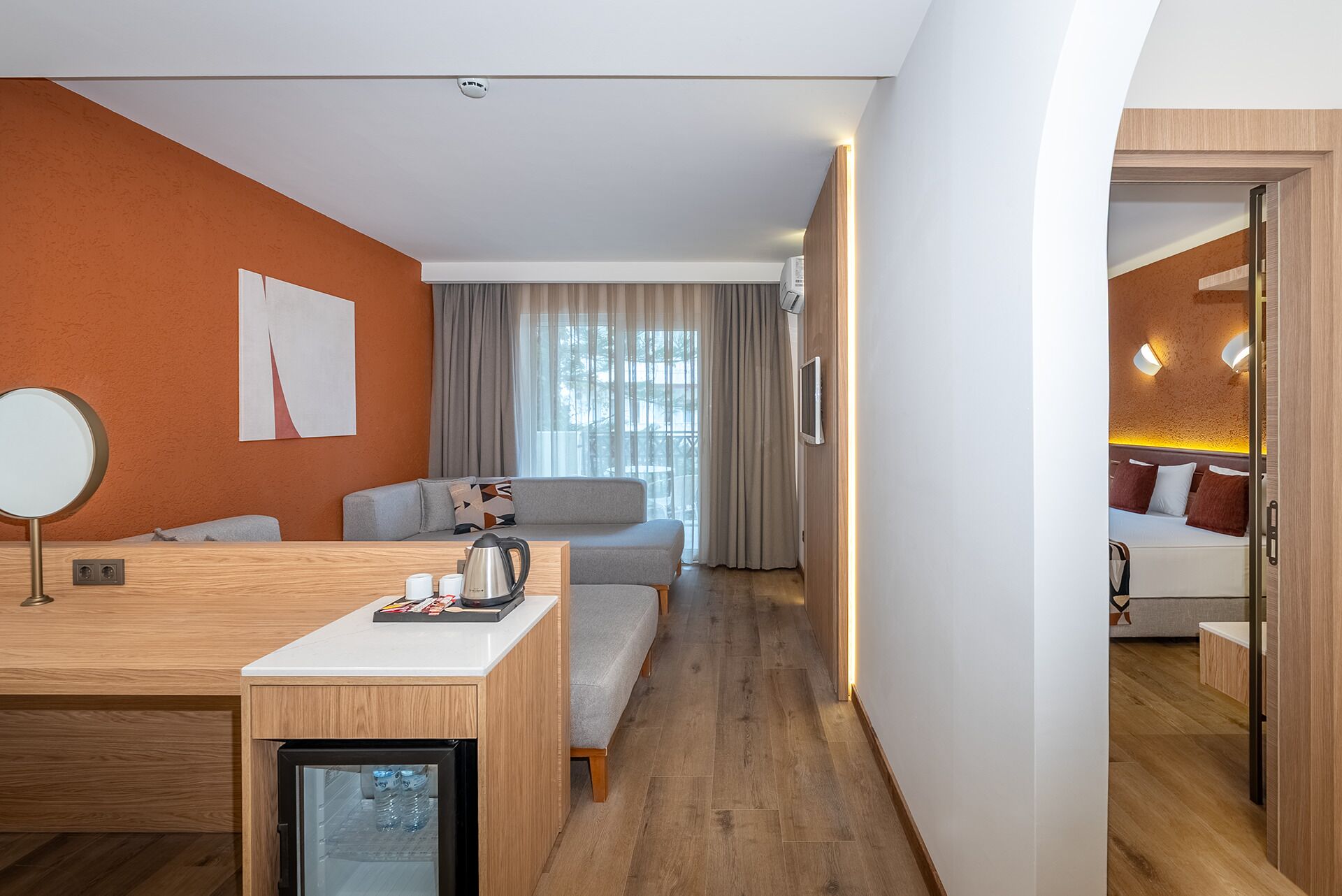 A modern hotel room with a wooden desk, a sofa, a bed, and a small kitchenette.