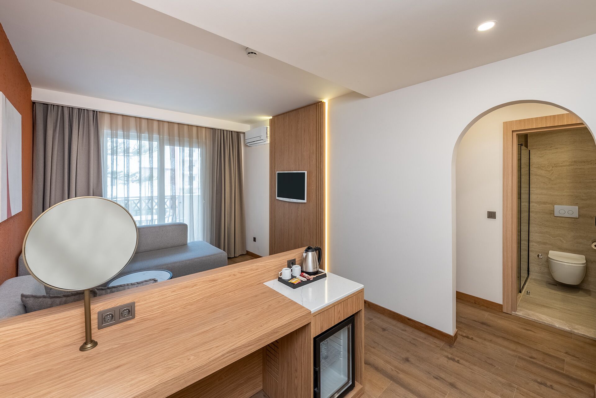 A modern hotel room with a wooden desk, a large mirror, a flat-screen TV, and a glass-enclosed shower.