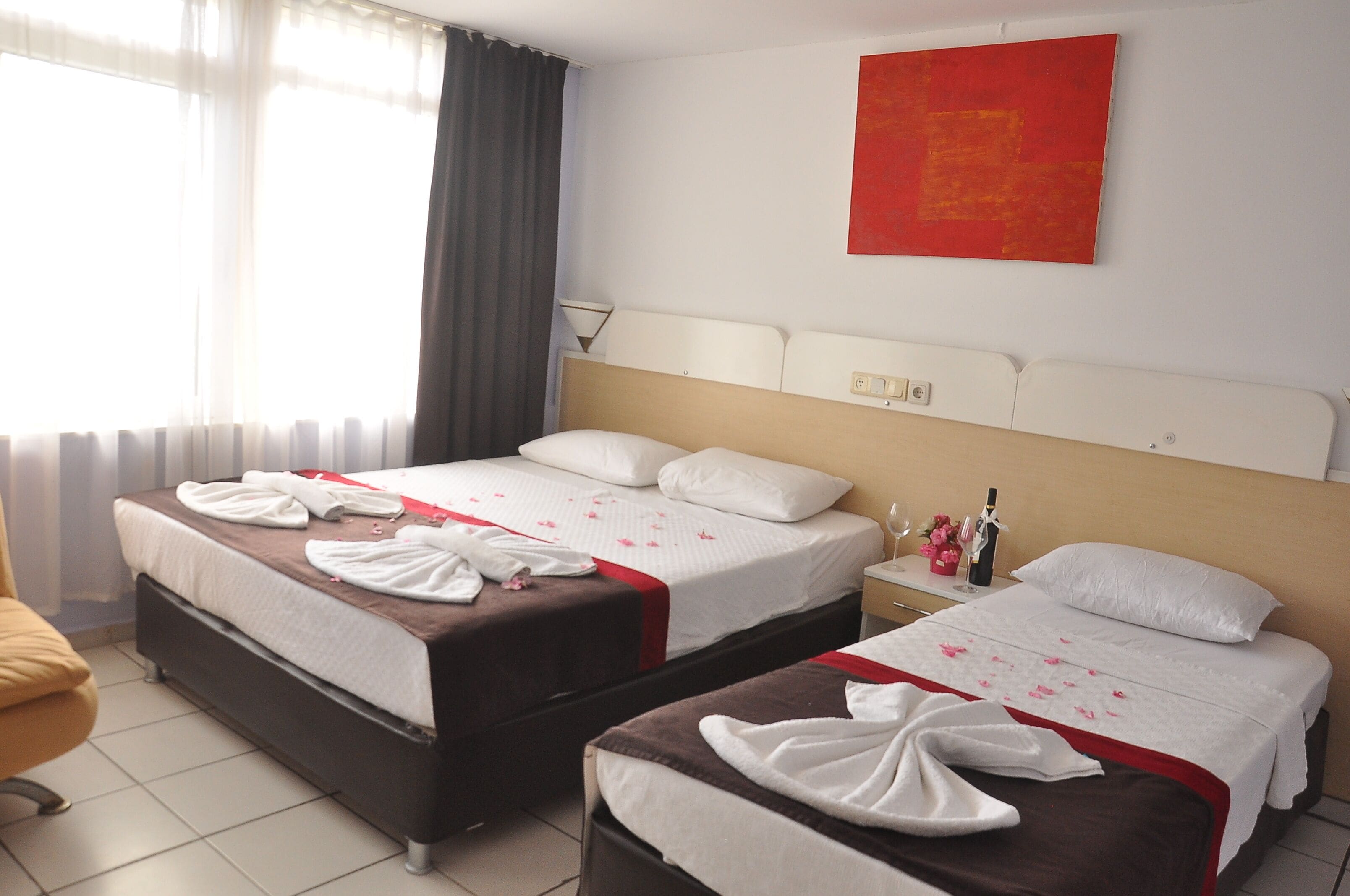 Een hotelkamer met twee bedden, elk voorzien van een witte spreidek en rode accenten, een abstract schilderij in rode tinten aan de muur, een nachtkastje met een lamp, en een raam met dunne gordijnen.