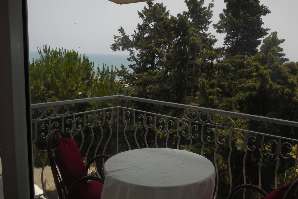 A balcony with a round table and red chairs, overlooking a view of trees and the sea.