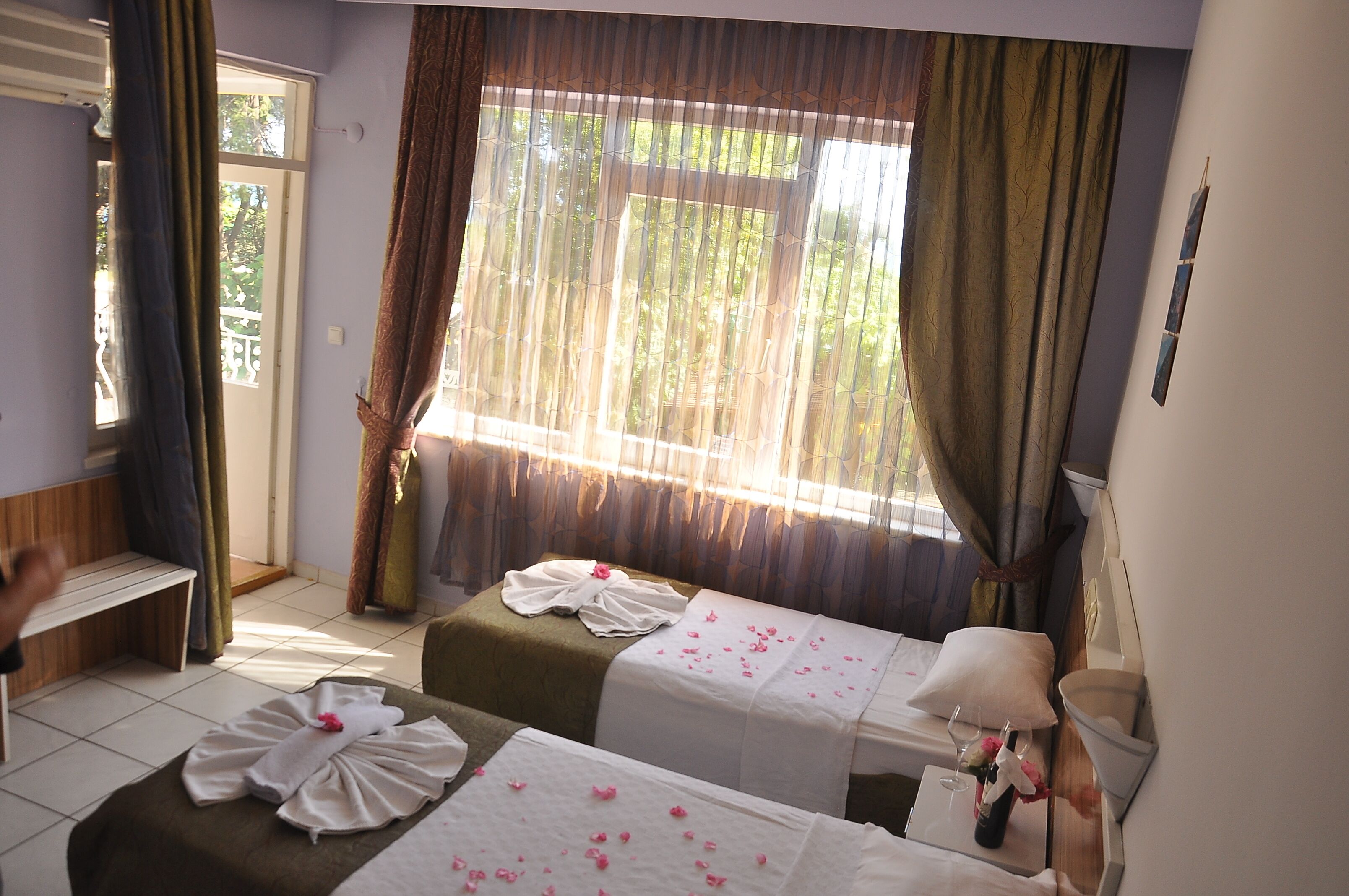 A hotel room with two beds, a bench, a window with curtains, and a bedside table with a lamp and a vase of flowers.