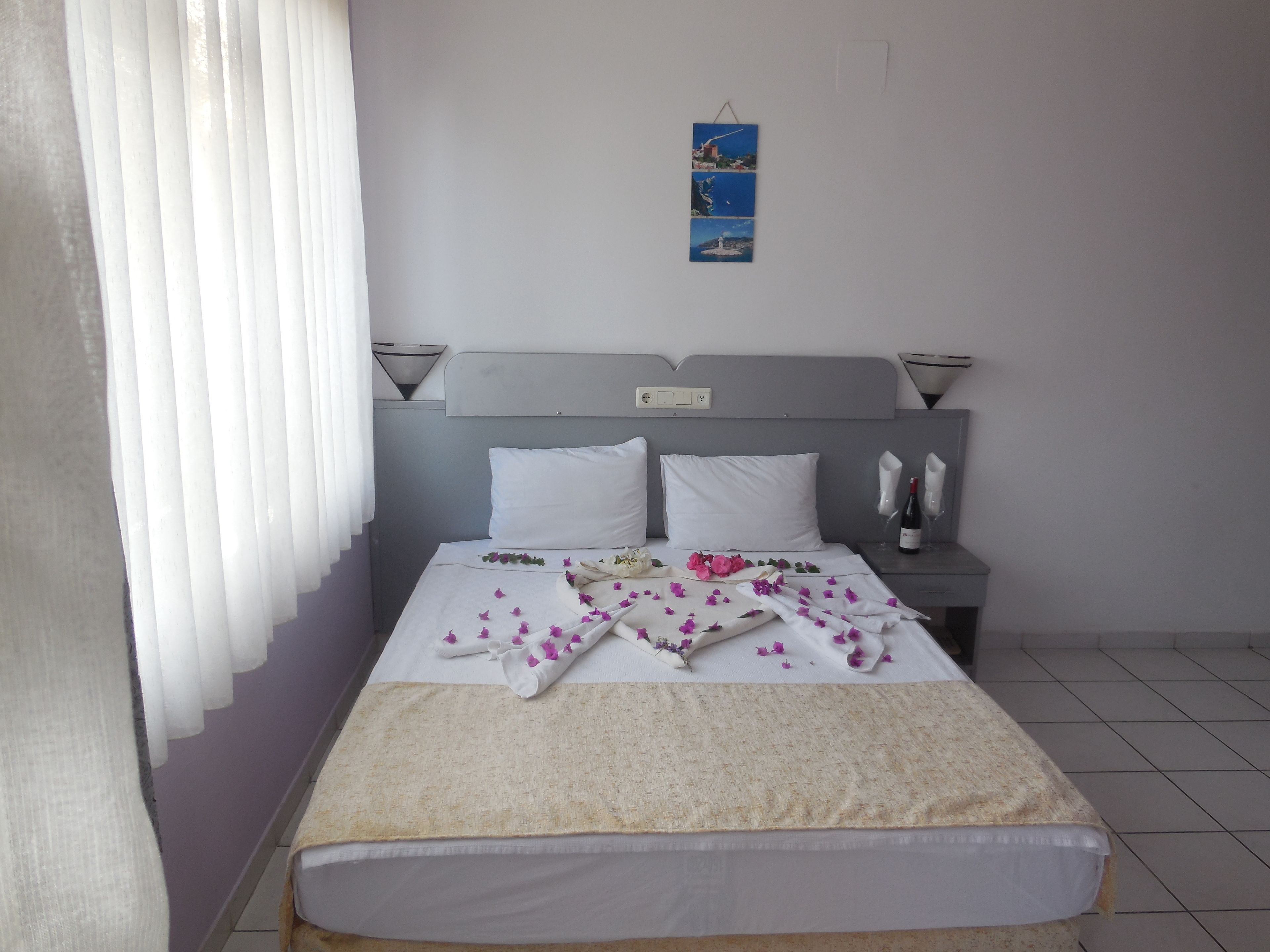 Una cama bien hecha con sábanas blancas y una manta color beige, una mesita de noche con una botella de vino y copas, y un cuadro colgado en la pared.