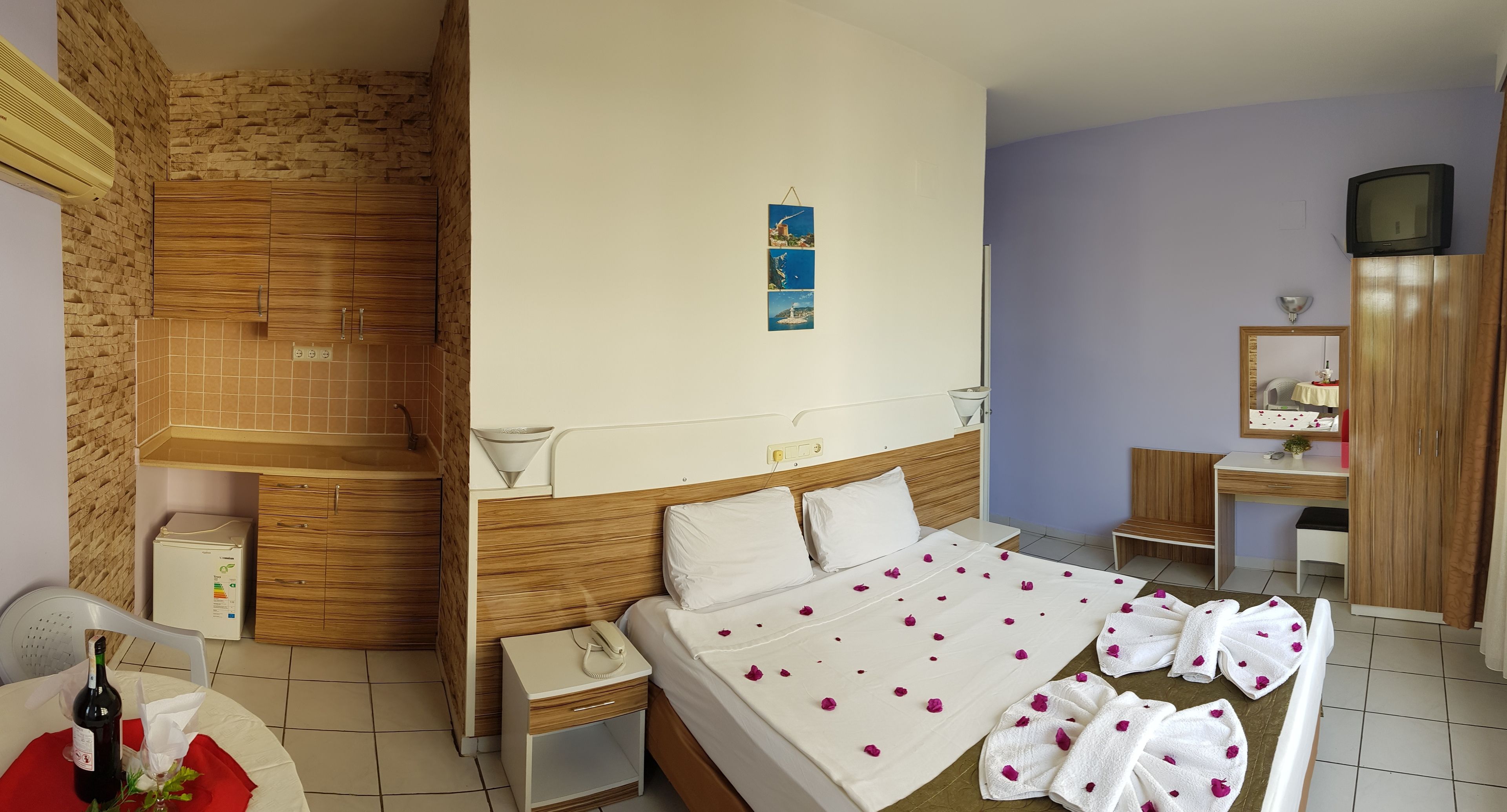 Quarto de hotel com uma cama, mesa de cabeceira com telefone, pequena escrivaninha com abajur, televisão em suporte de madeira e parede de tijolos.