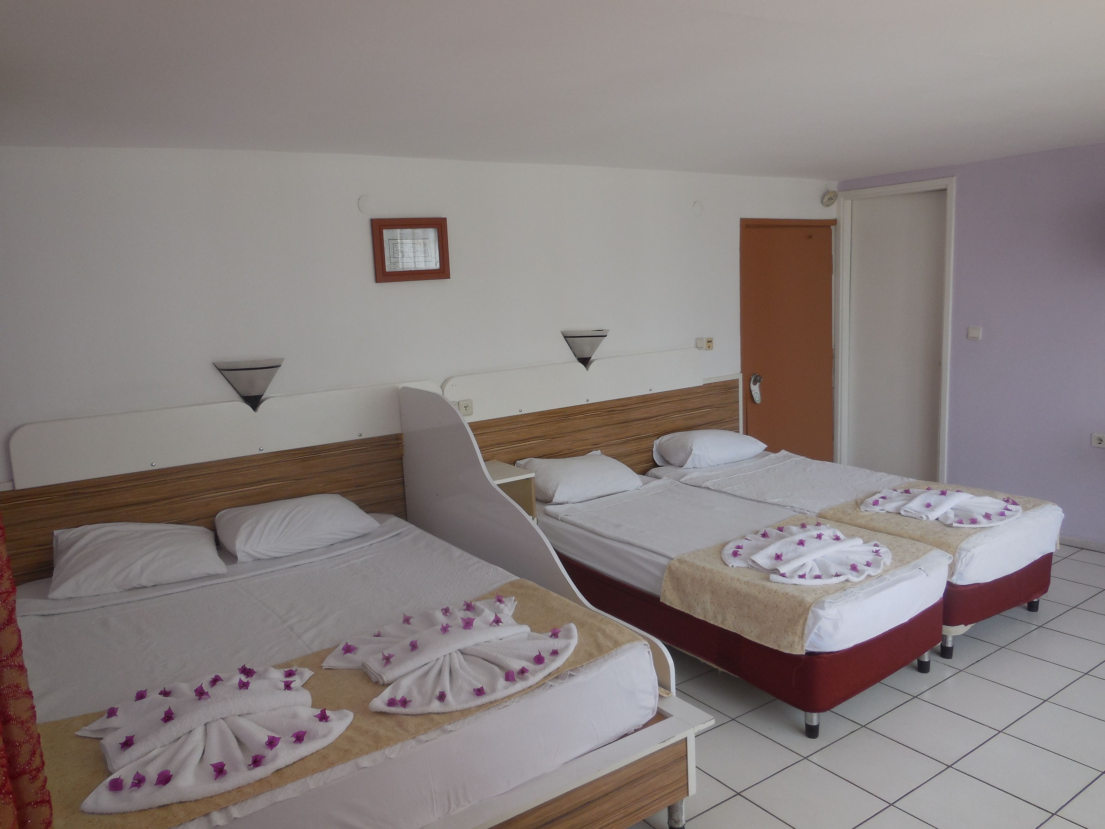 A hotel room with two beds, each with a wooden headboard, white bedding, and towels with purple flowers. There is a small framed picture on the wall and a door to the right.