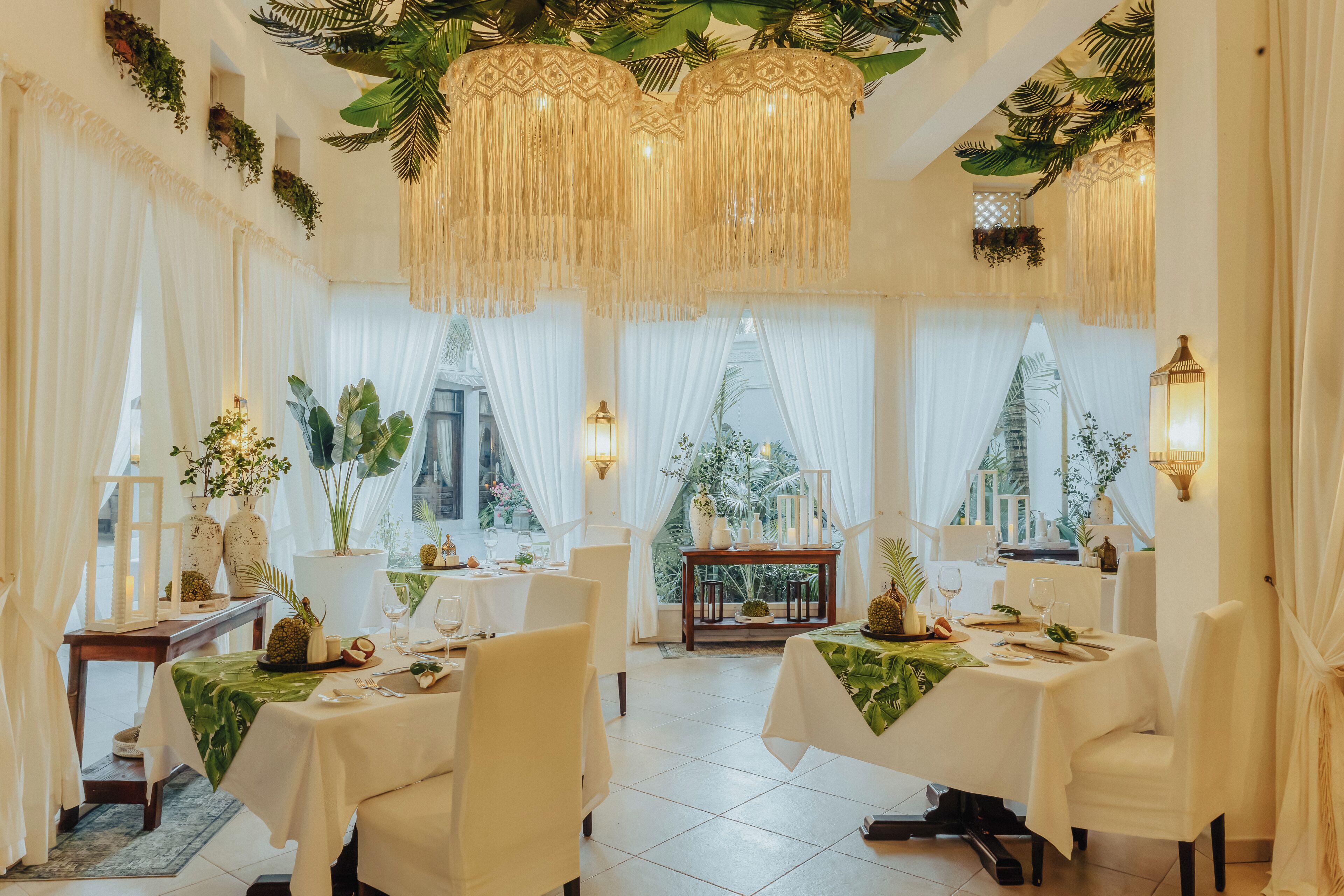 A dining area with tables set for two, white drapes, hanging plants, and a view of the outdoors.