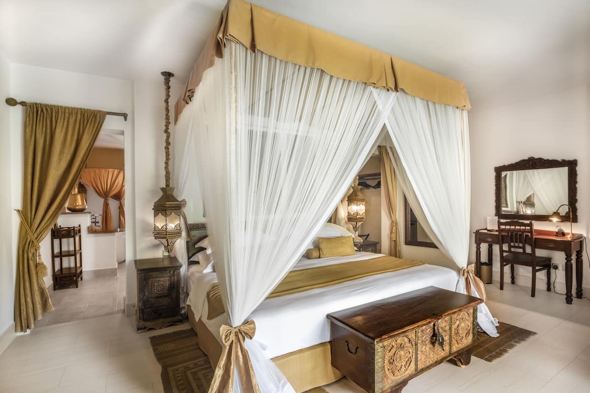 A four-poster bed with a canopy, a wooden nightstand, a desk with a chair, and a mirror on the wall.