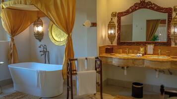 Une salle de bain avec une baignoire sur pieds, une chaise en bois avec une serviette, un grand miroir décoratif et deux appliques murales.