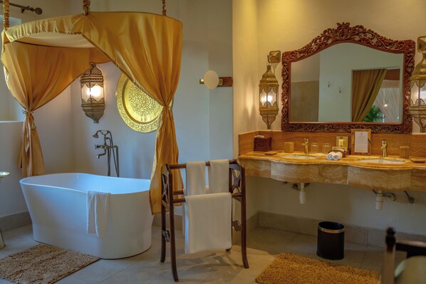 Un baño con una bañera independiente, una silla de madera con una toalla, un espejo grande y ornamentado, y dos luces fijadas a la pared.