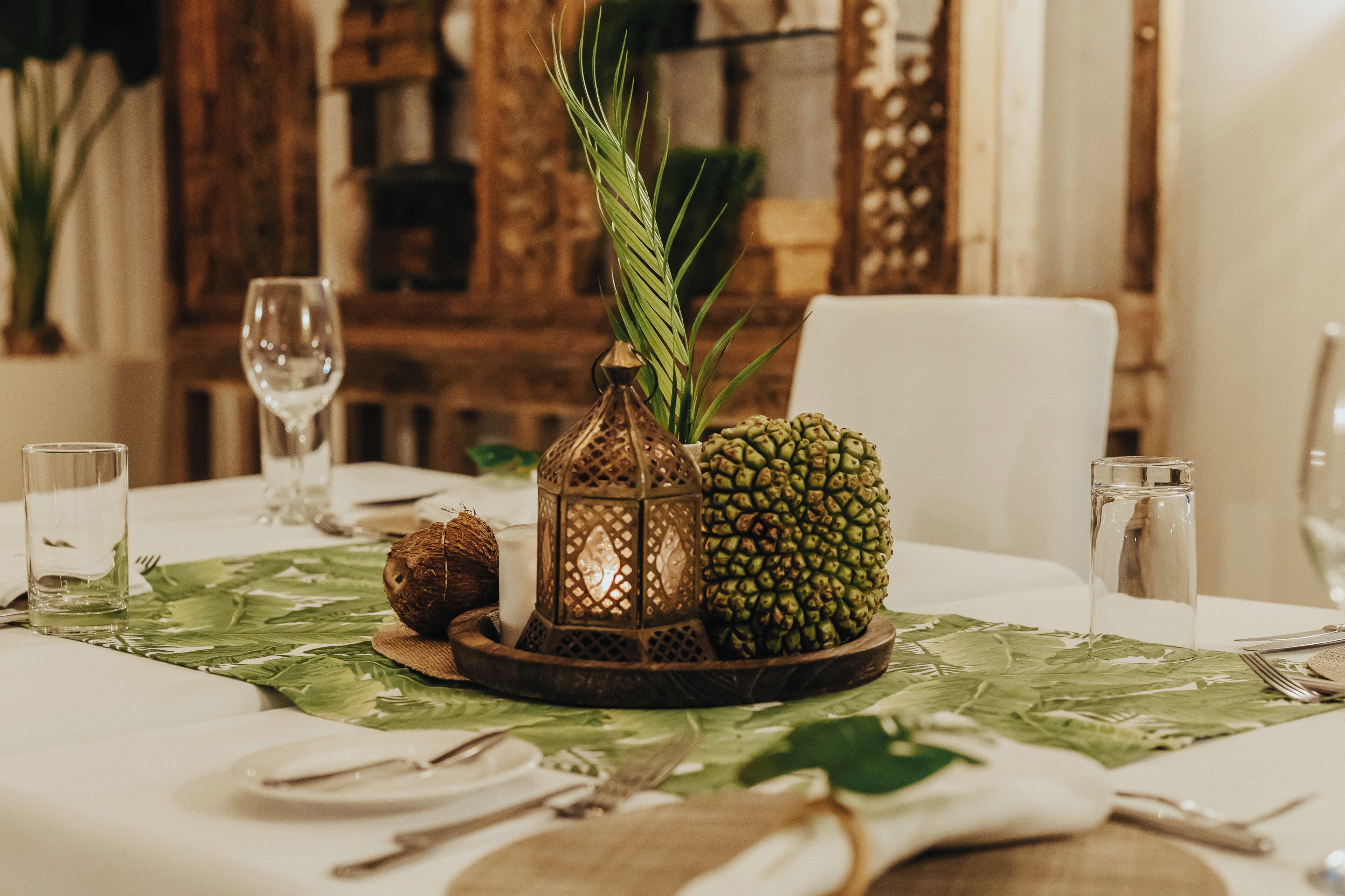 A dining table with a green leaf-patterned tablecloth, a pineapple centerpiece, a coconut, a lantern, and wine glasses.