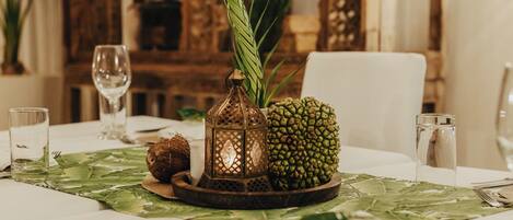 Una mesa de comedor con mantel estampado de hojas verdes, un centro de mesa con piña, un coco, un farol y copas de vino.