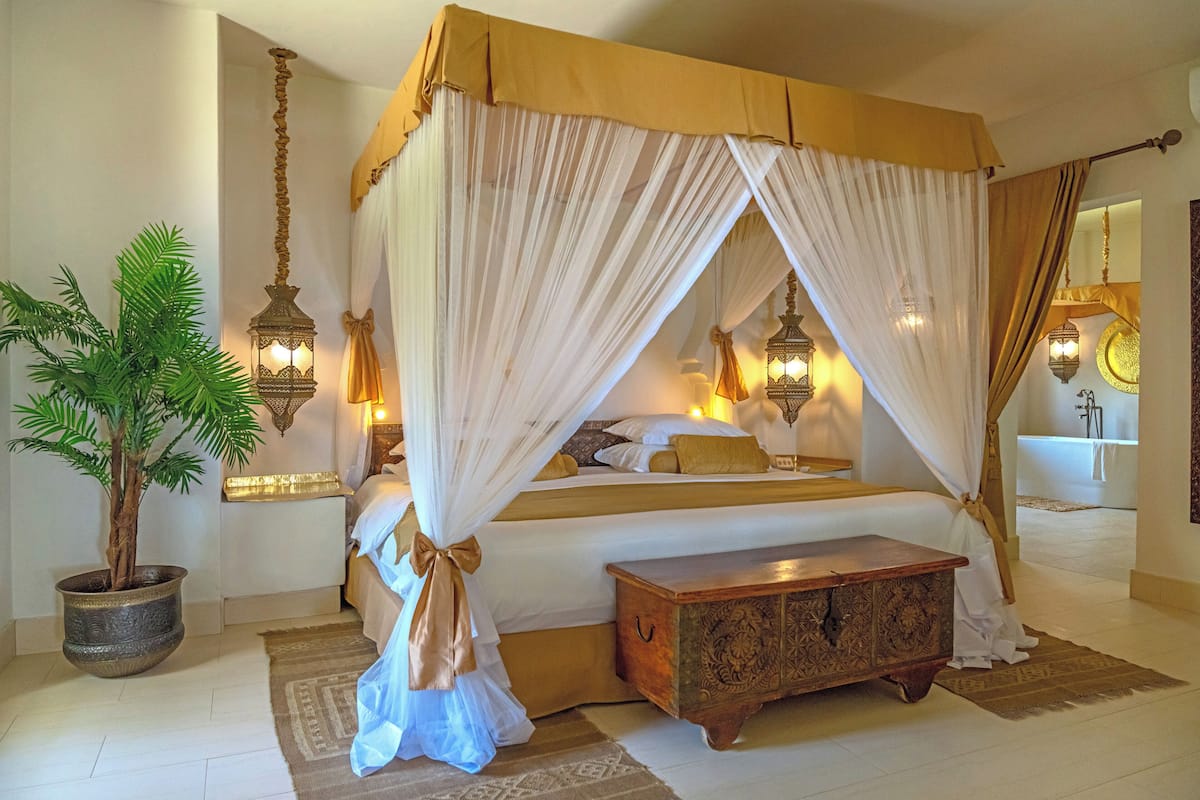 A bedroom with a four-poster bed, a nightstand, a potted plant, and a wooden chest of drawers.