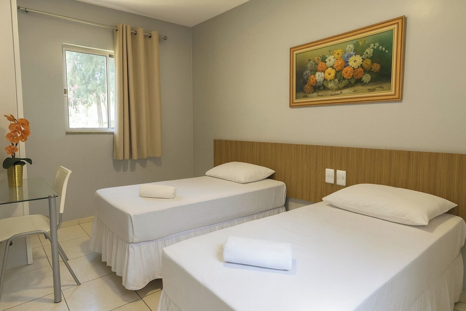 A hotel room with two beds, a dining table, a chair, a window with curtains, and a painting on the wall.