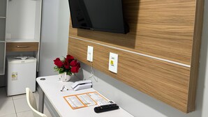 Een hotelkamer met een wandgemonteerde flatscreen televisie, een wit bureau met een vaas rode rozen, een kleine koelkast en een stopcontact op de muur.