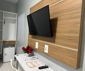 Une chambre d’hôtel équipée d’un téléviseur à écran plat fixé au mur, d’un bureau blanc orné d’un vase de roses rouges, d’un petit réfrigérateur et d’une prise électrique murale.