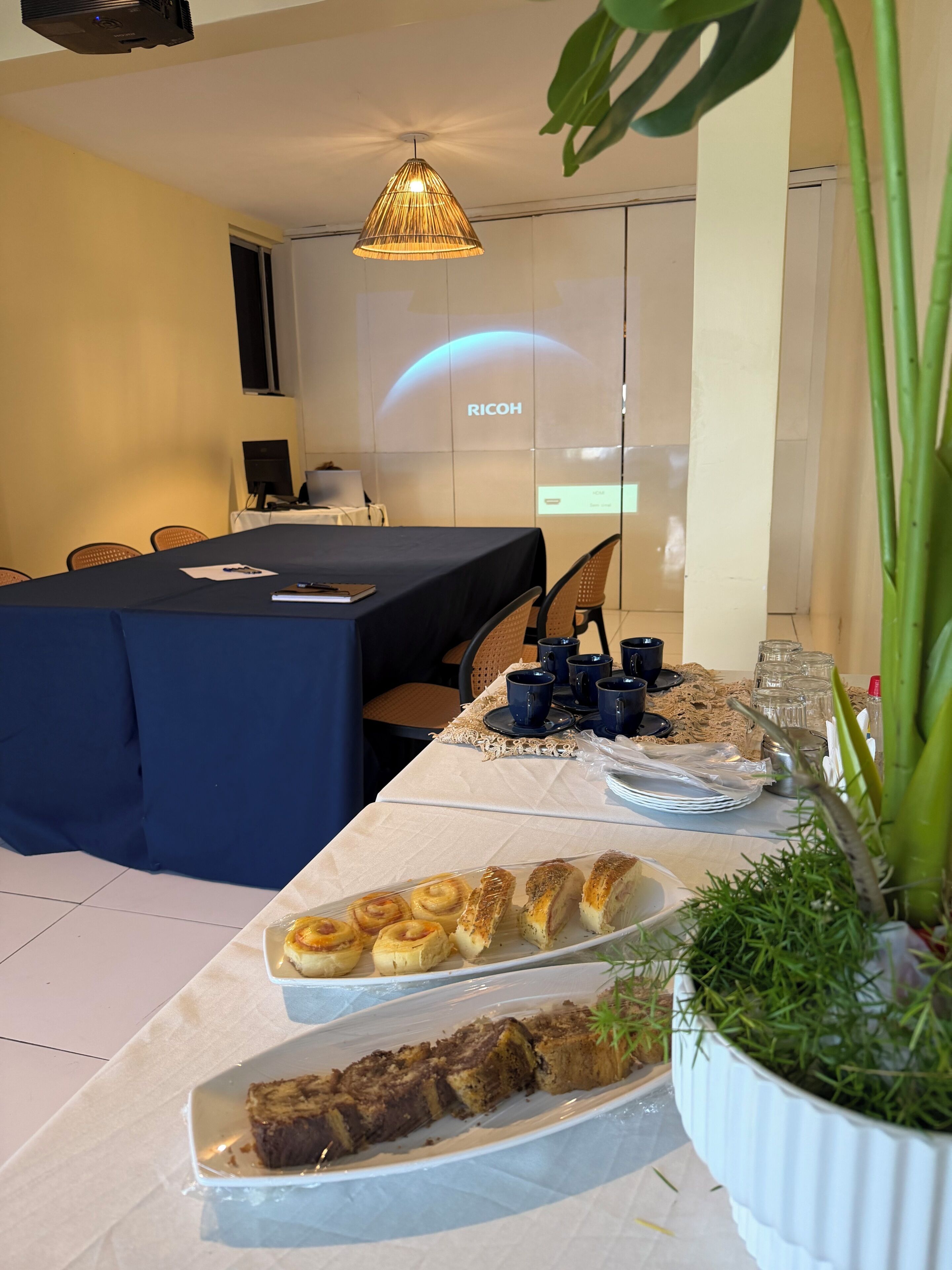 Un salón de conferencias con una pantalla de proyector mostrando 'RICOH', una mesa con platos de pasteles y una planta en maceta.