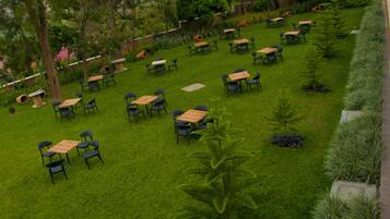 Un espacio al aire libre con varias mesas y sillas dispuestas sobre un césped, rodeado de árboles y plantas.