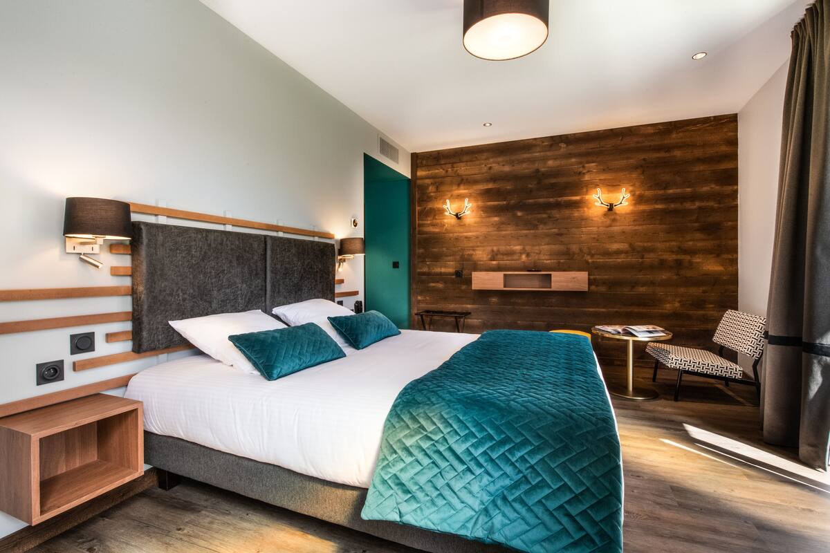 Una habitación de hotel moderna con una cama grande, un panel de madera decorativo, una pequeña mesa con una silla y una lámpara fijada a la pared.