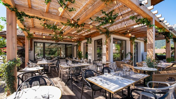 Un patio al aire libre con mesas y sillas de madera, cubierto por una pérgola con plantas colgantes y luces de cuerda.