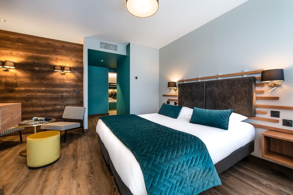 Una habitación de hotel moderna con una cama grande, una pared de acento de madera, una zona de estar con un reposapiés amarillo y vistas al baño.