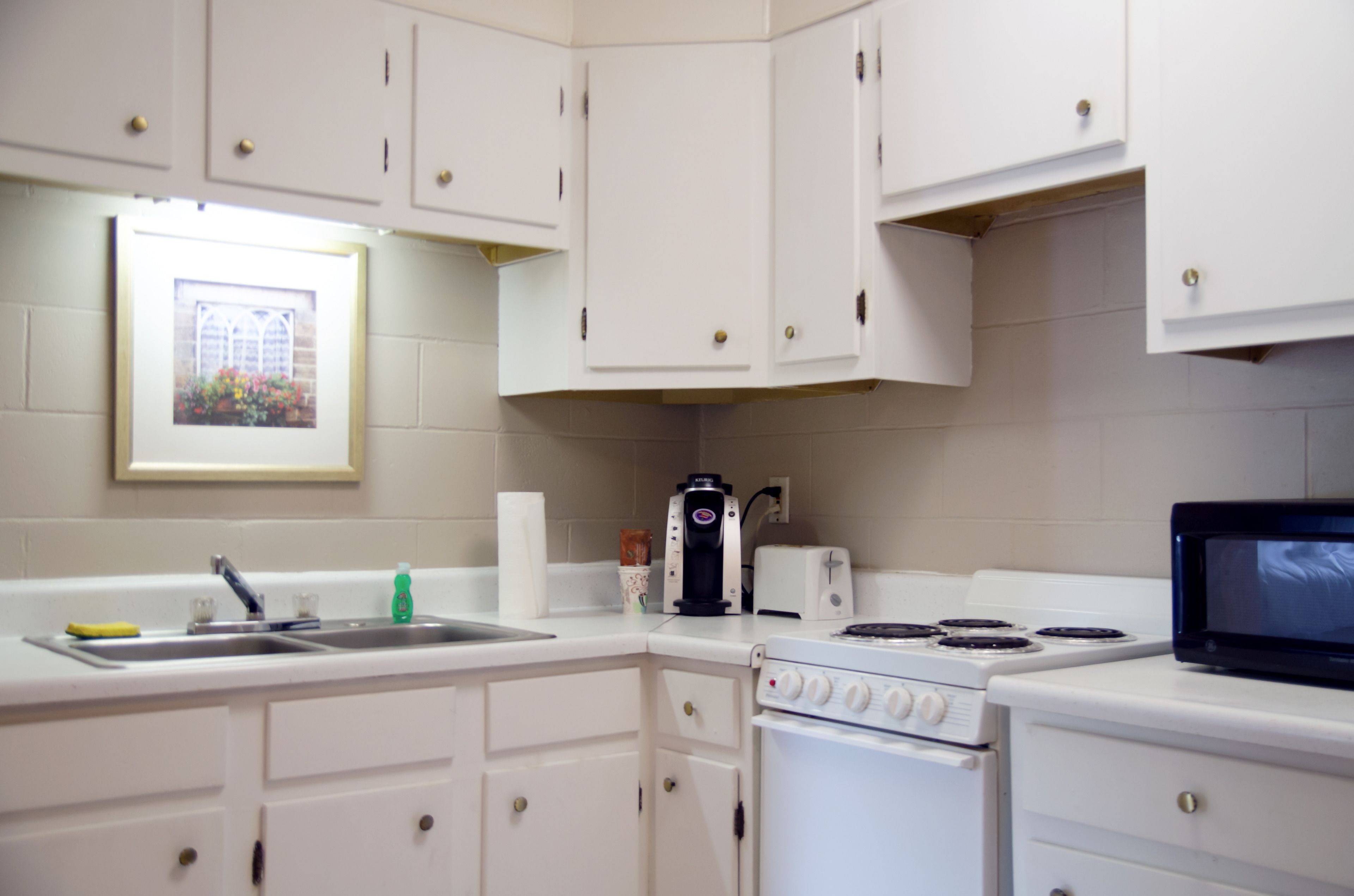 A kitchen with white cabinets, a stove, a microwave, a coffee maker, and a framed picture on the wall.