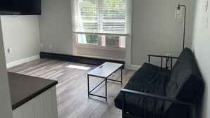 A living room with a black leather sofa, a small table, and a window with blinds. - Seabreeze Motel (Old Orchard Beach)