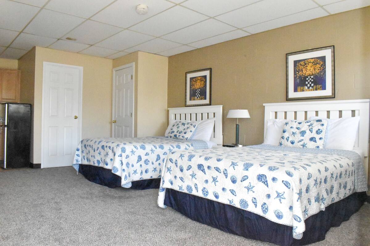 A hotel room with two beds, a door, a refrigerator, and two wall-mounted lamps.