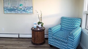 A room with a patterned blue sofa, a round wooden side table with a vase and a small box, and a painting of sailboats on the wall. - Seabreeze Motel (Old Orchard Beach)