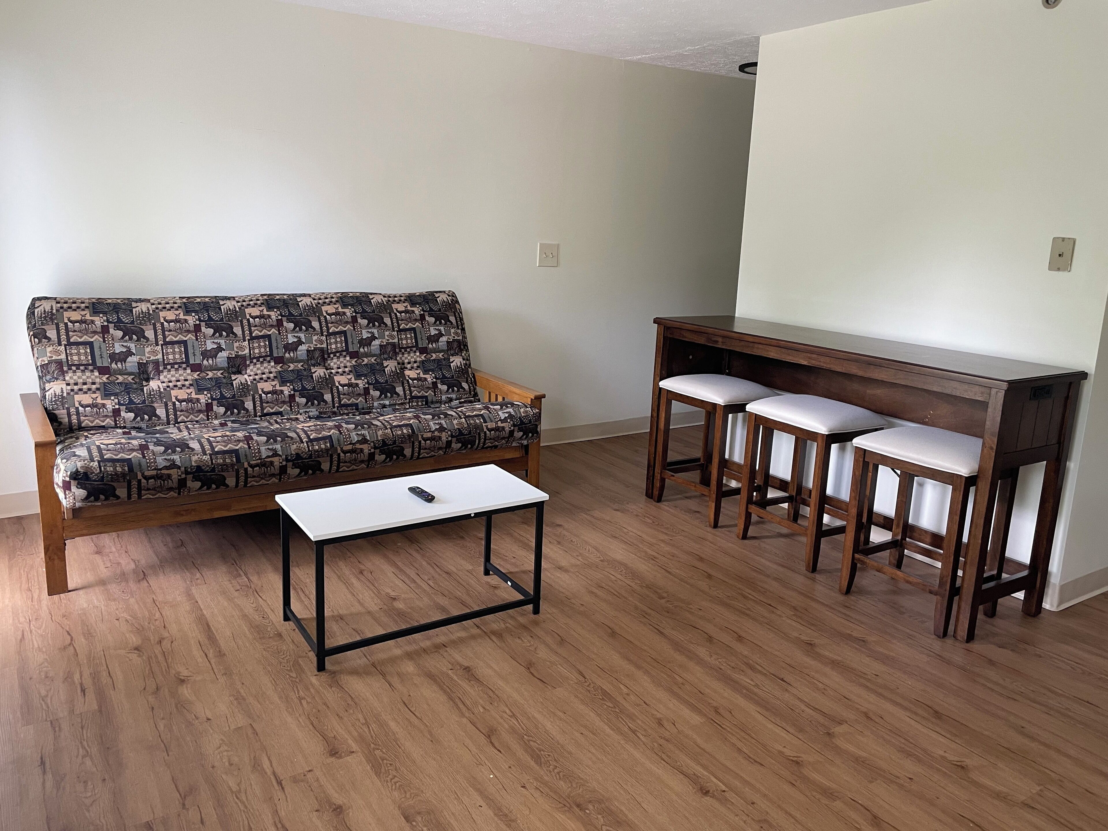 A living room with a patterned sofa, a small white table, and a wooden bar with two stools.