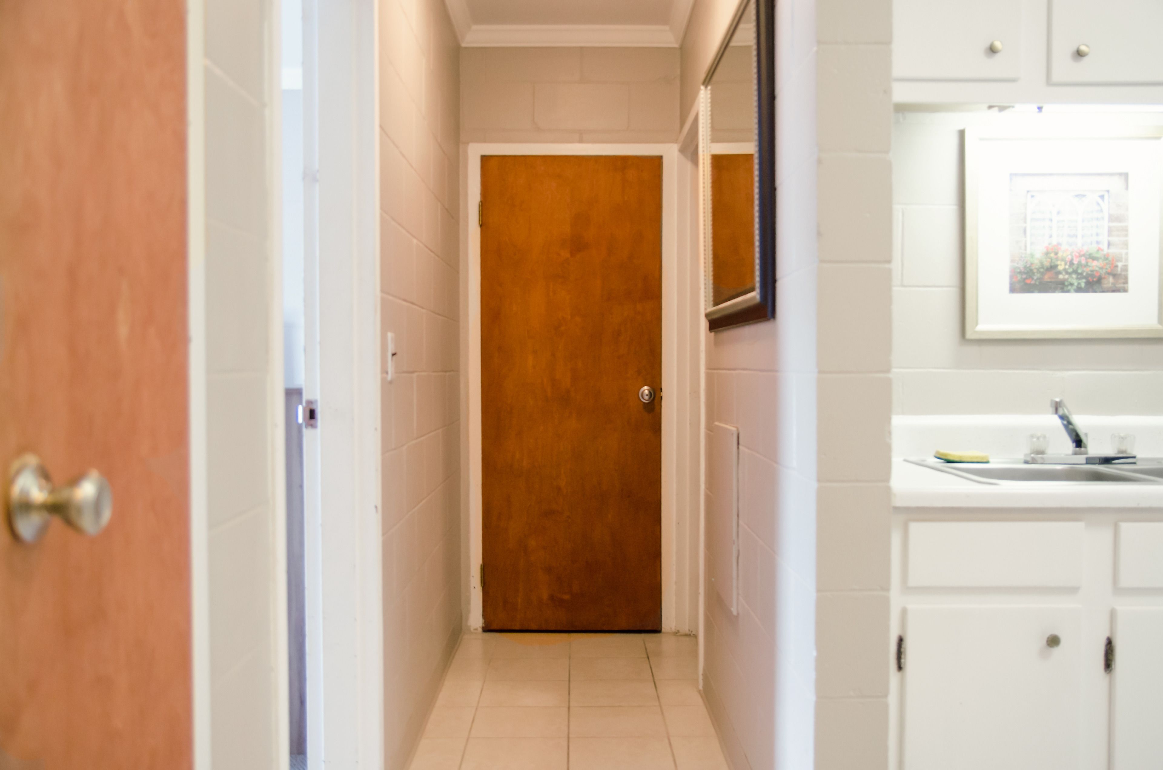 A narrow hallway with a wooden door, a white tiled floor, and a white bathroom with a sink and mirror.