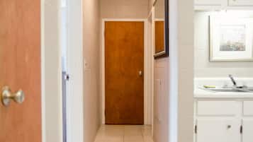 A narrow hallway with a wooden door, a white tiled floor, and a white bathroom with a sink and mirror.