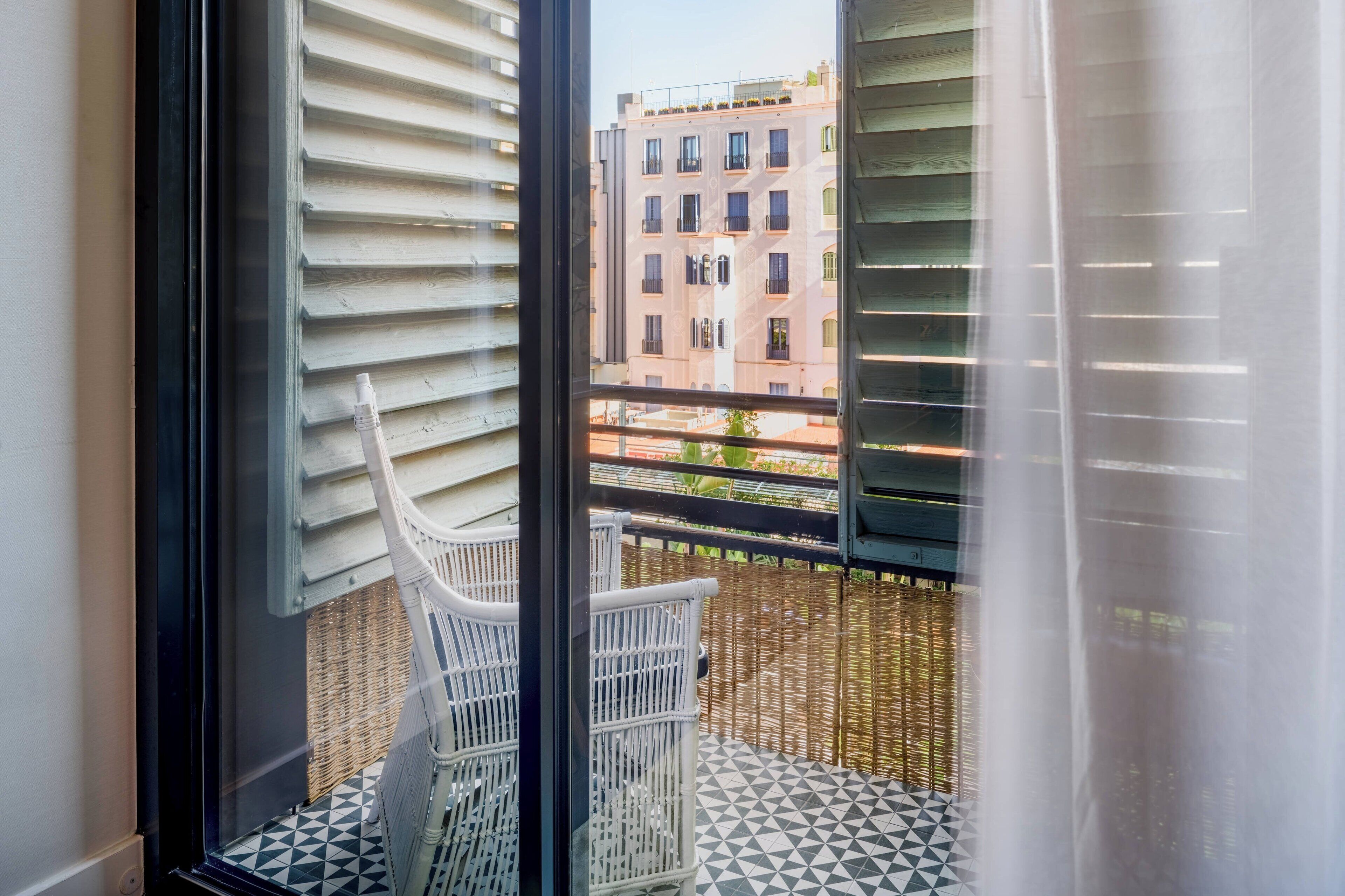 Un balcon doté d’un sol à motifs géométriques, de meubles en osier et offrant une vue sur un immeuble à multiples fenêtres.