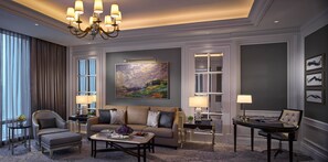 A well-appointed living room with a sofa, armchairs, a coffee table, and a dining table. There is a large painting on the wall, a chandelier, and a view through a window.