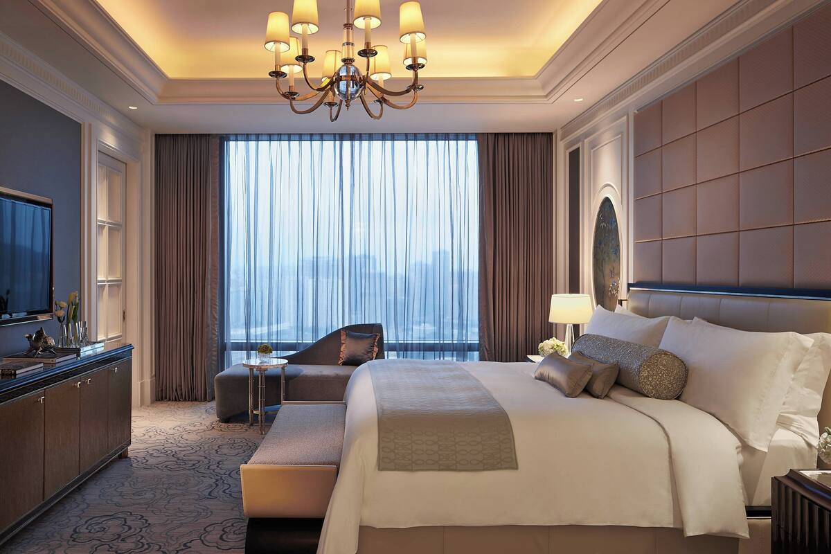 A spacious hotel room with a large bed, a sitting area, a television, and a view of the city through large windows.