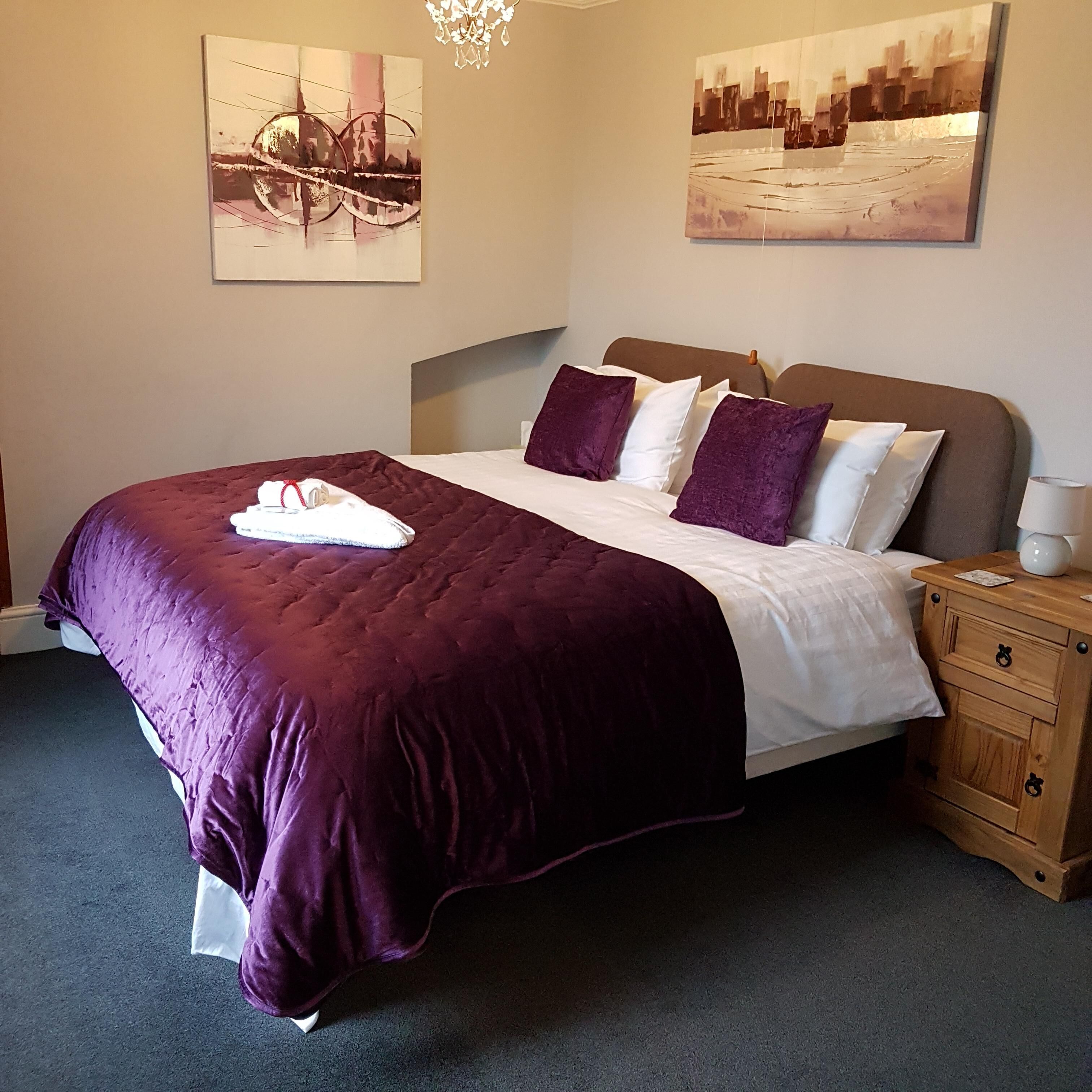 A neatly made bed with a purple bedspread and white linens, a wooden nightstand with a lamp, and two wall-mounted paintings.