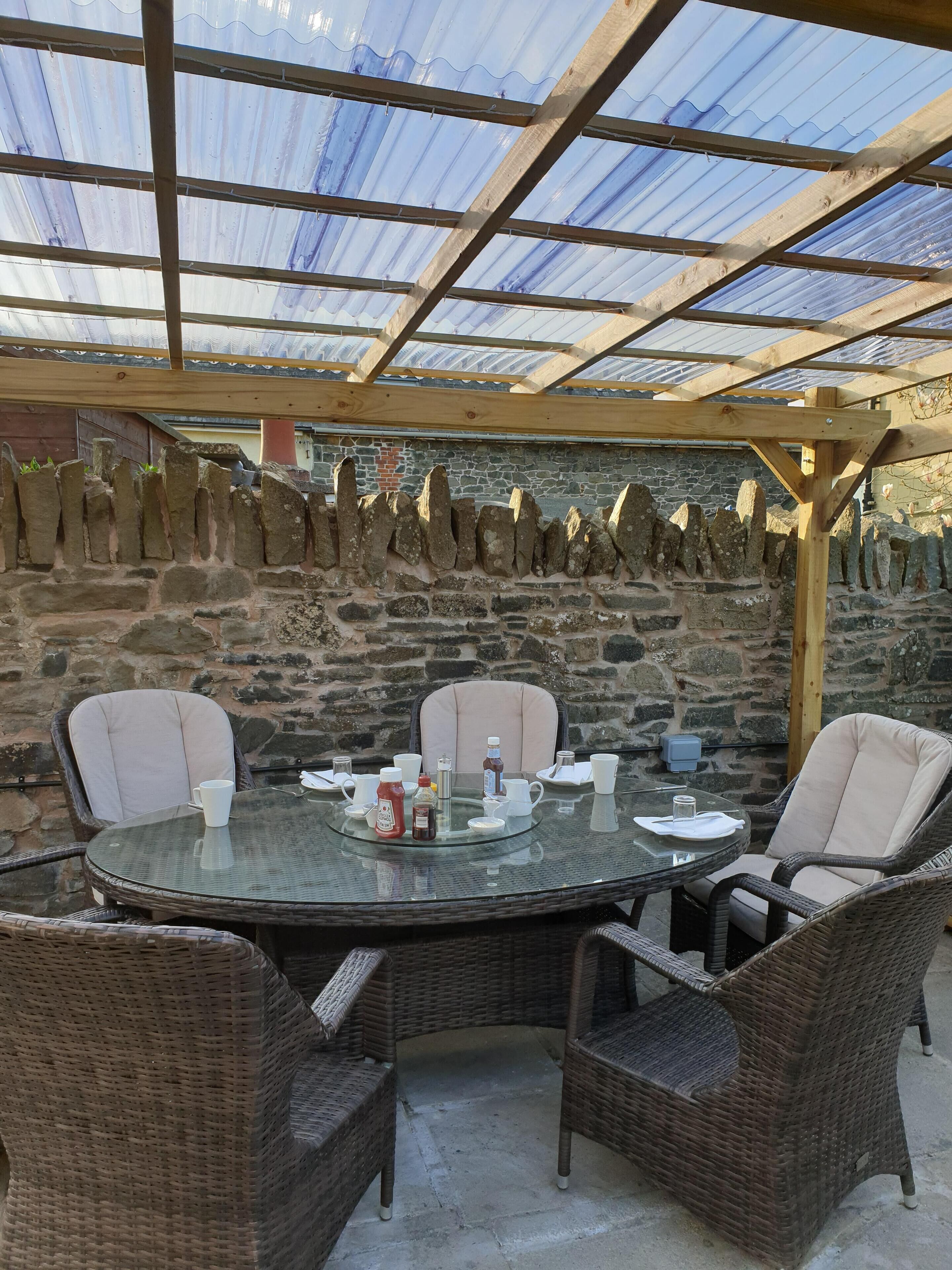 A covered outdoor dining area with a stone wall backdrop, wicker chairs, and a glass table set for four.